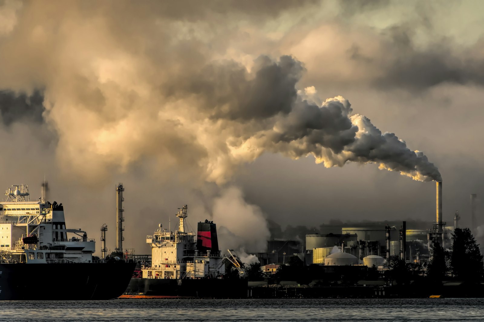 A coastal refinery and tank farm at dusk with a single stack venting a long plume across the sky, two tankers moored at the jetty in front