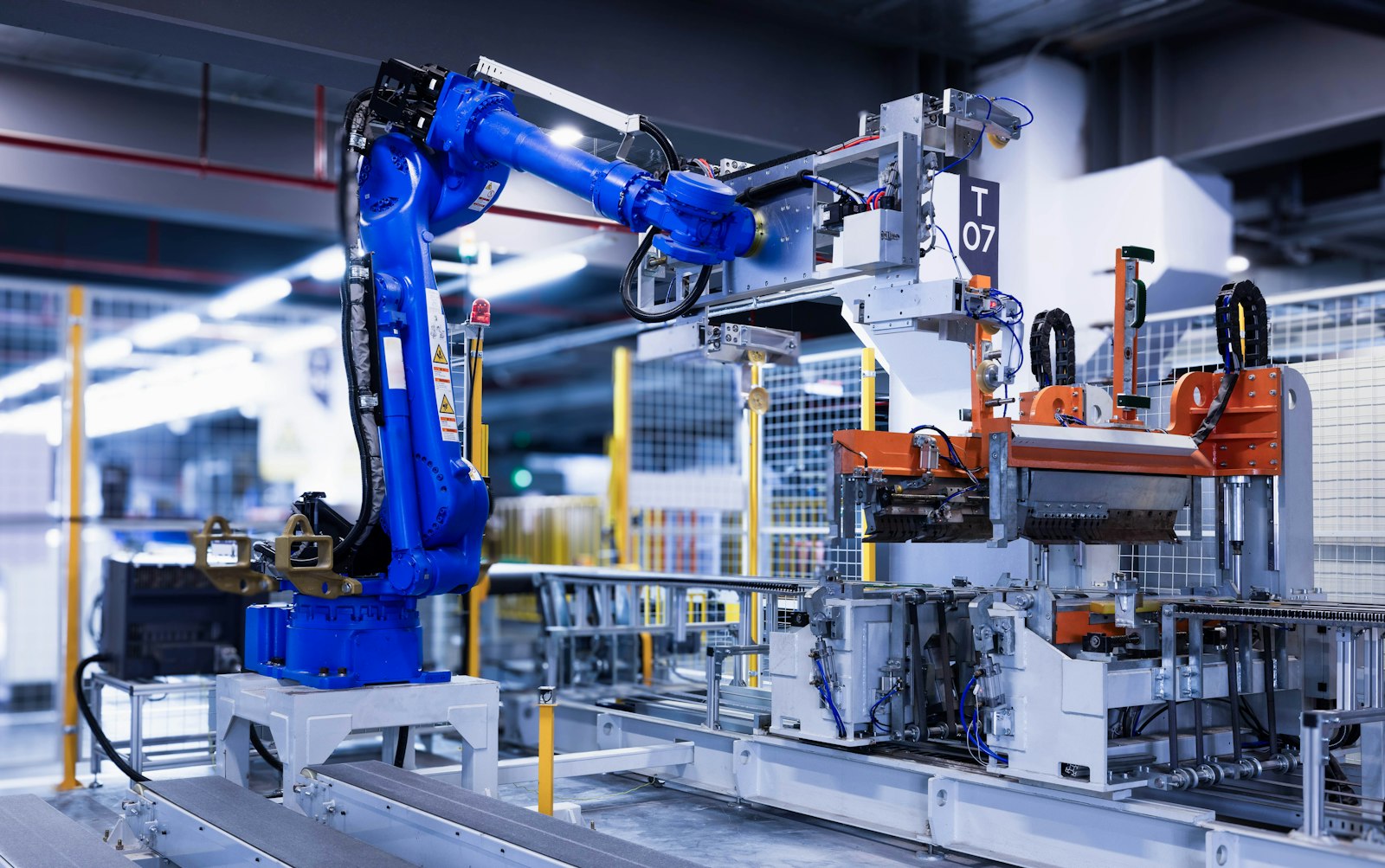 A blue six-axis industrial robot arm on a rail beside an automated assembly cell in a brightly lit factory