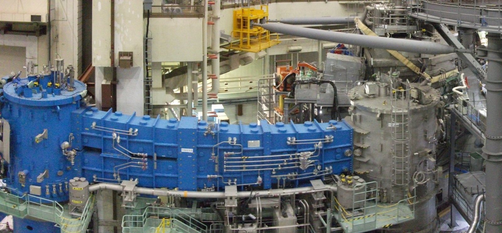 The JT-60SA fusion experiment hall in Naka, Ibaraki, during assembly in 2016: large blue equipment modules and a grey vessel with ladders and walkways, surrounded by pipework and steel gantries