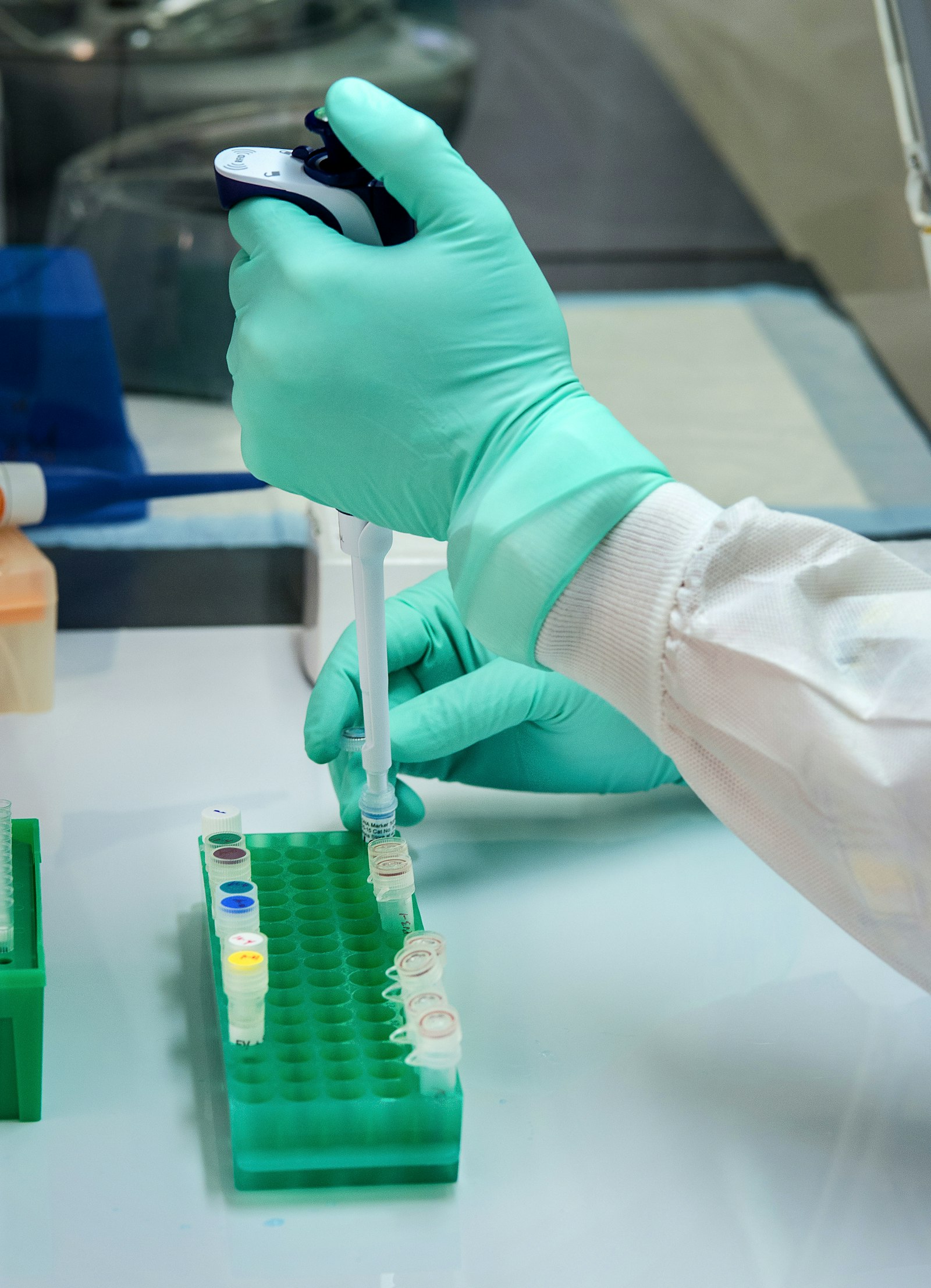 A gloved hand in a white lab coat pipetting into a green rack of labelled microtubes on a clean bench