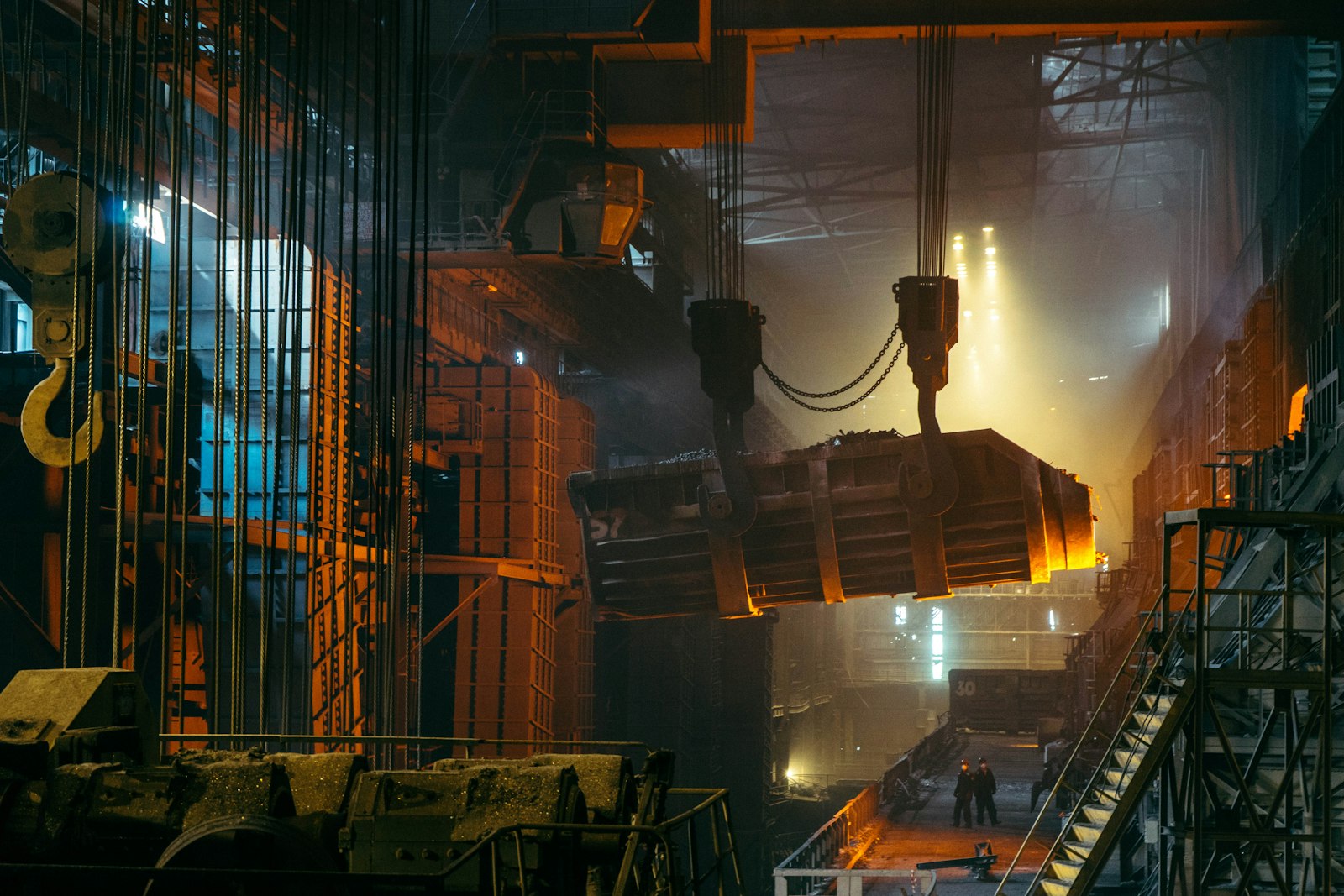 Overhead crane carrying a glowing charge ladle through the haze of a steelworks casting bay, workers visible on the floor below